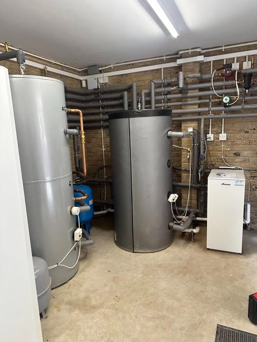 A plant room with two grey hot water cylinders, a white floor-standing boiler, an expansion vessel and lagged pipework on a brick wall